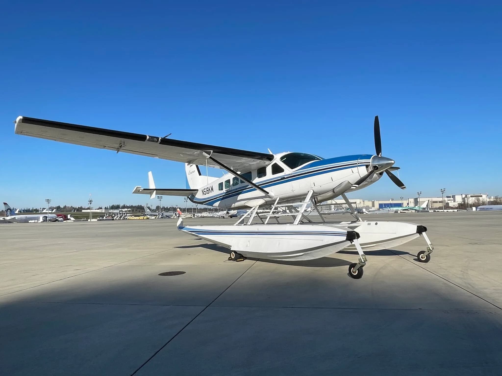 A white and blue Cessna Caravan floatplane (N518KM) with floats parked on an airfield under a clear blue sky.