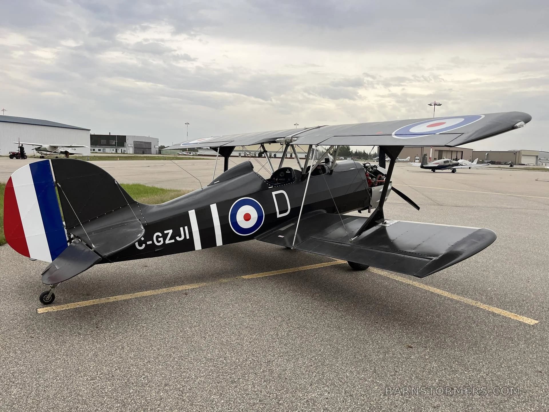 A black biplane with a silver engine cowling and British roundel markings, parked on an airport tarmac under an overcast sky.