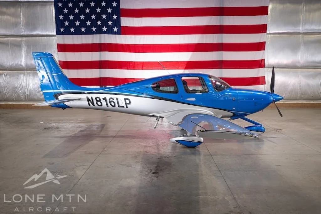 A blue and silver Cirrus SR22 aircraft, registration N816LP, parked in a hangar with an American flag in the background.