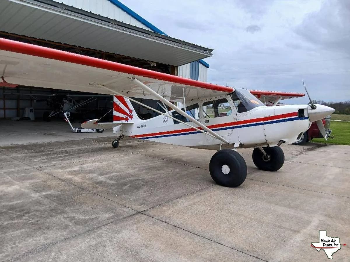 A white high-wing Maule aircraft with red and blue stripes and large bush tires, parked on a concrete apron next to a hangar.