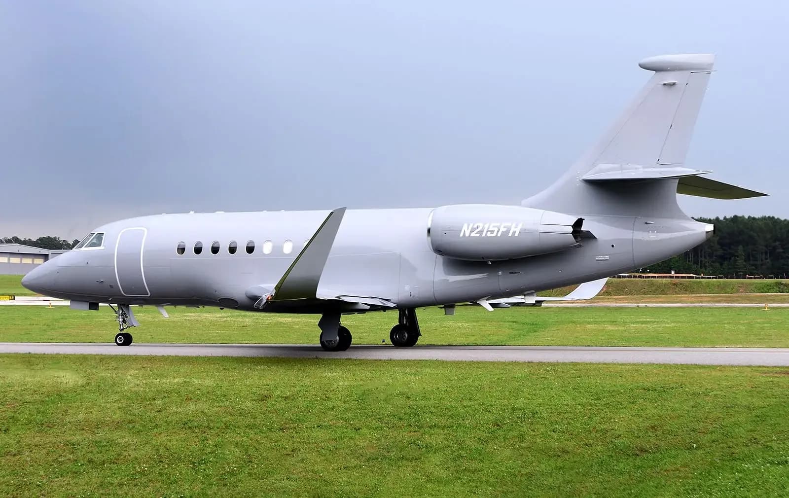 A side view of the Dassault Falcon 2000S business jet, registration N215FH, in matte grey, parked on an airfield with green grass.