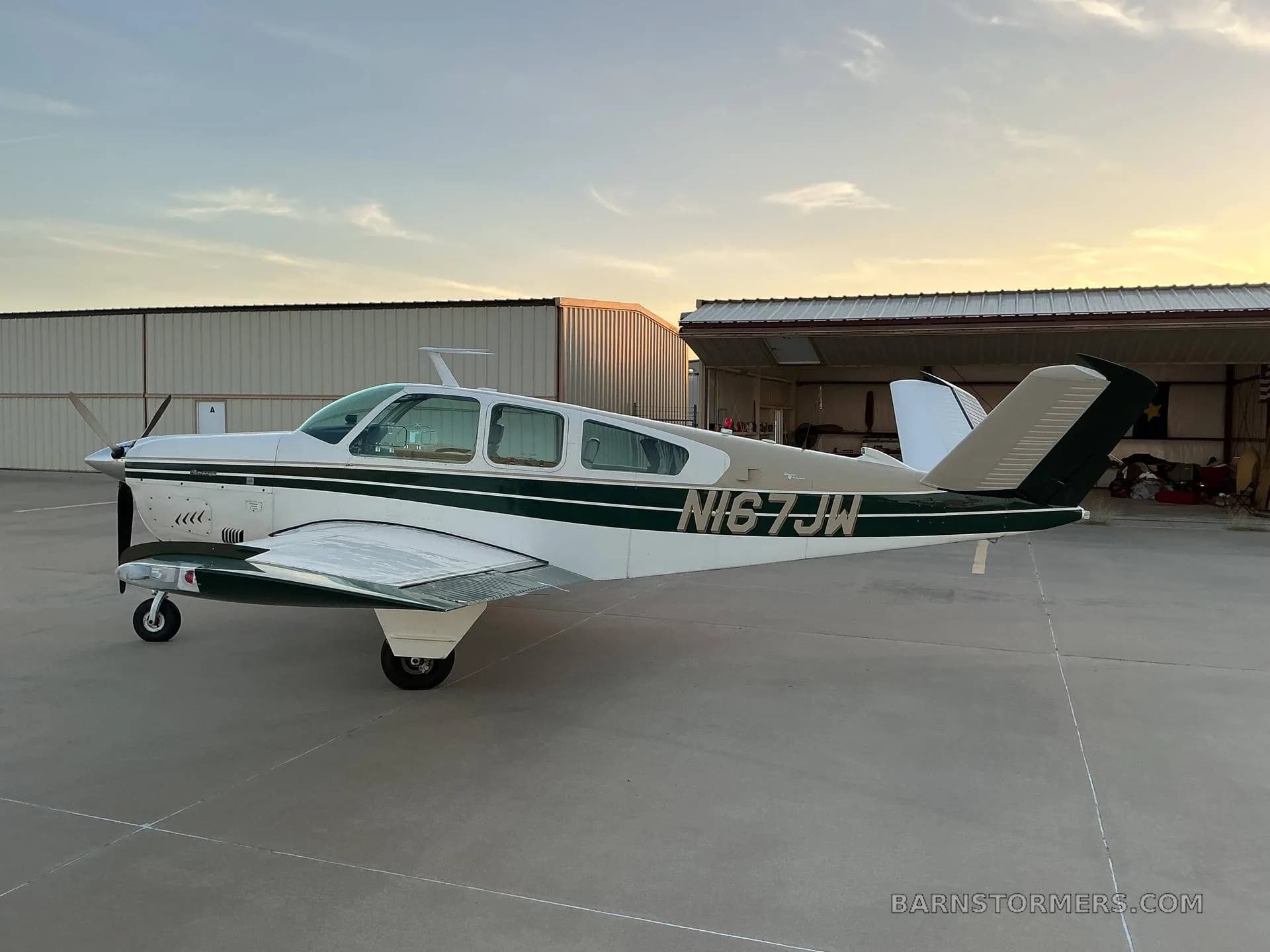 A white and dark green Beechcraft Bonanza (N167JW) parked on an airfield tarmac at sunset, with hangars in the background.
