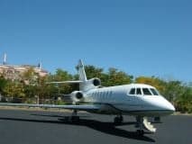 A white aircraft is parked on the tarmac in front of a building and trees.