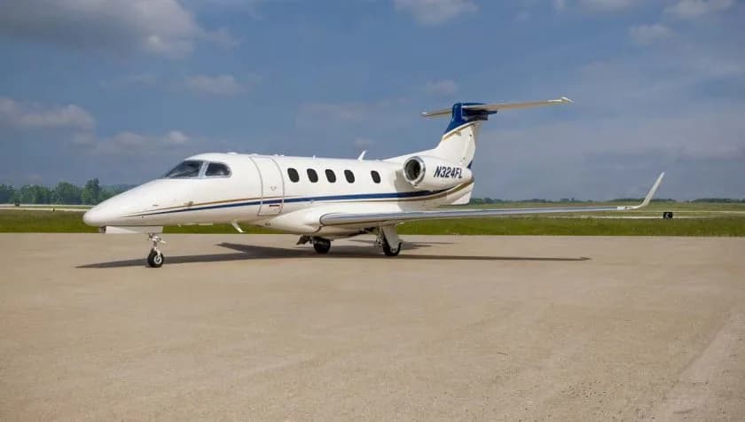 A white Embraer Phenom 300 private jet with blue and gold stripes on the tail and fuselage, parked on an airfield under a partly cloudy sky.
