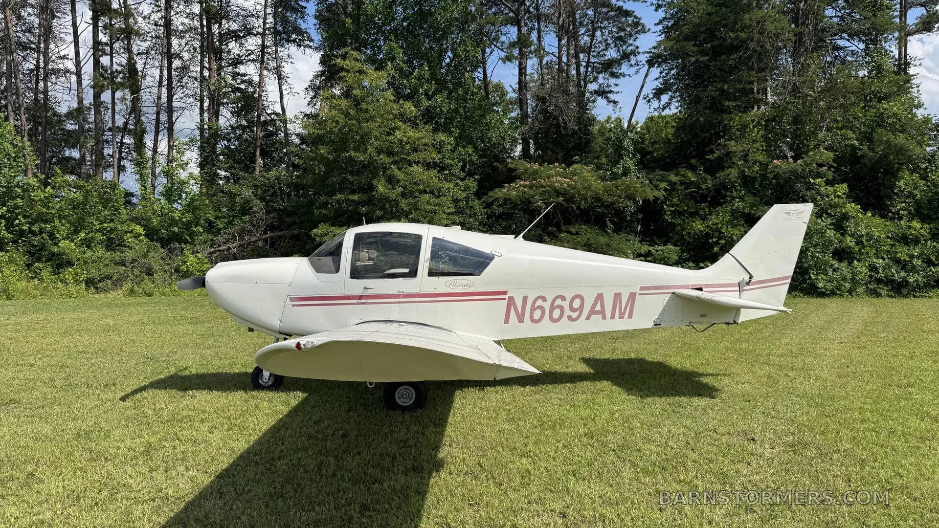A white single-engine propeller aircraft with red stripes, registration N669AM, parked on a grassy field with trees in the background, viewed from the side.