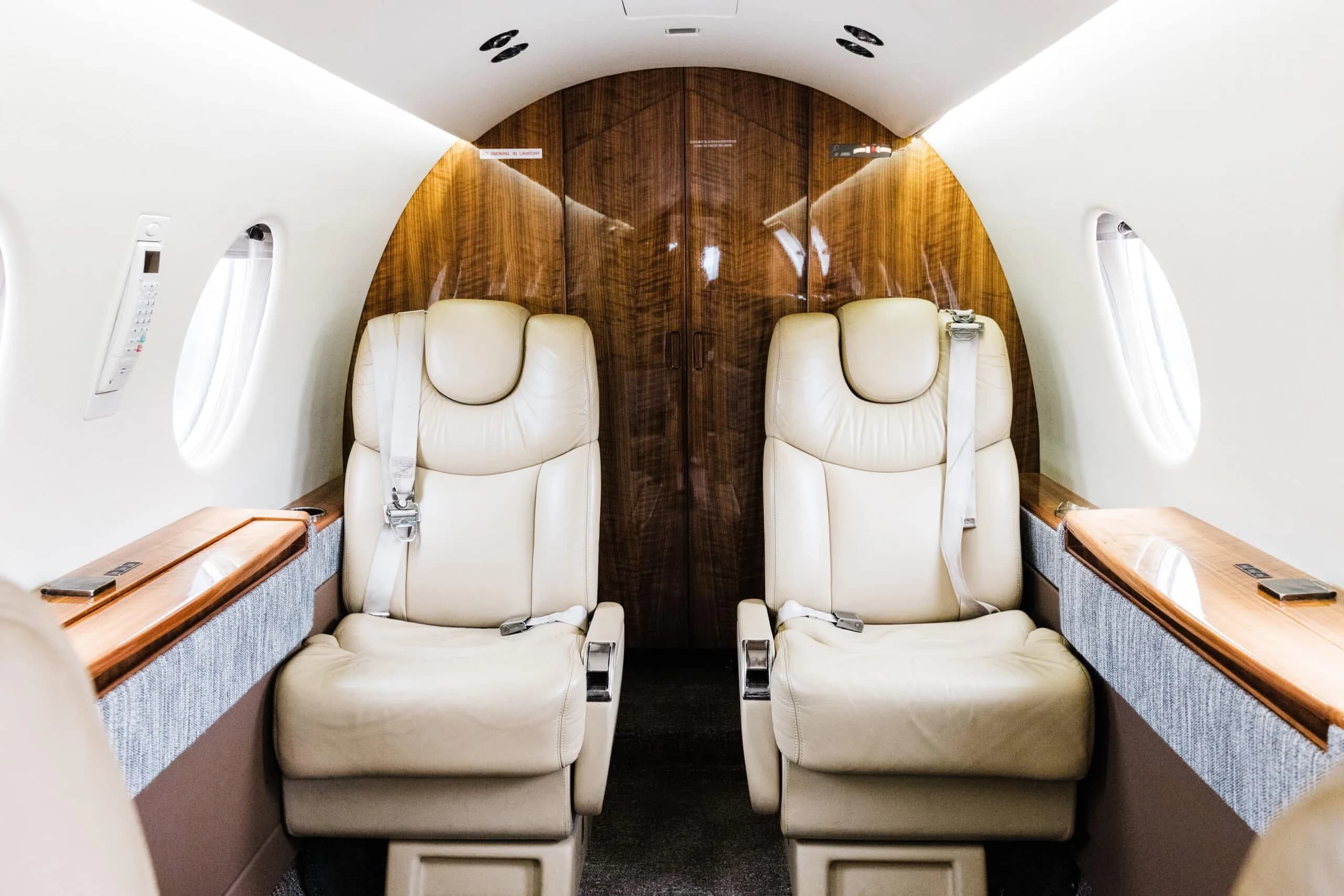 The private jet's cabin interior, showing two beige leather seats facing a wooden bulkhead.