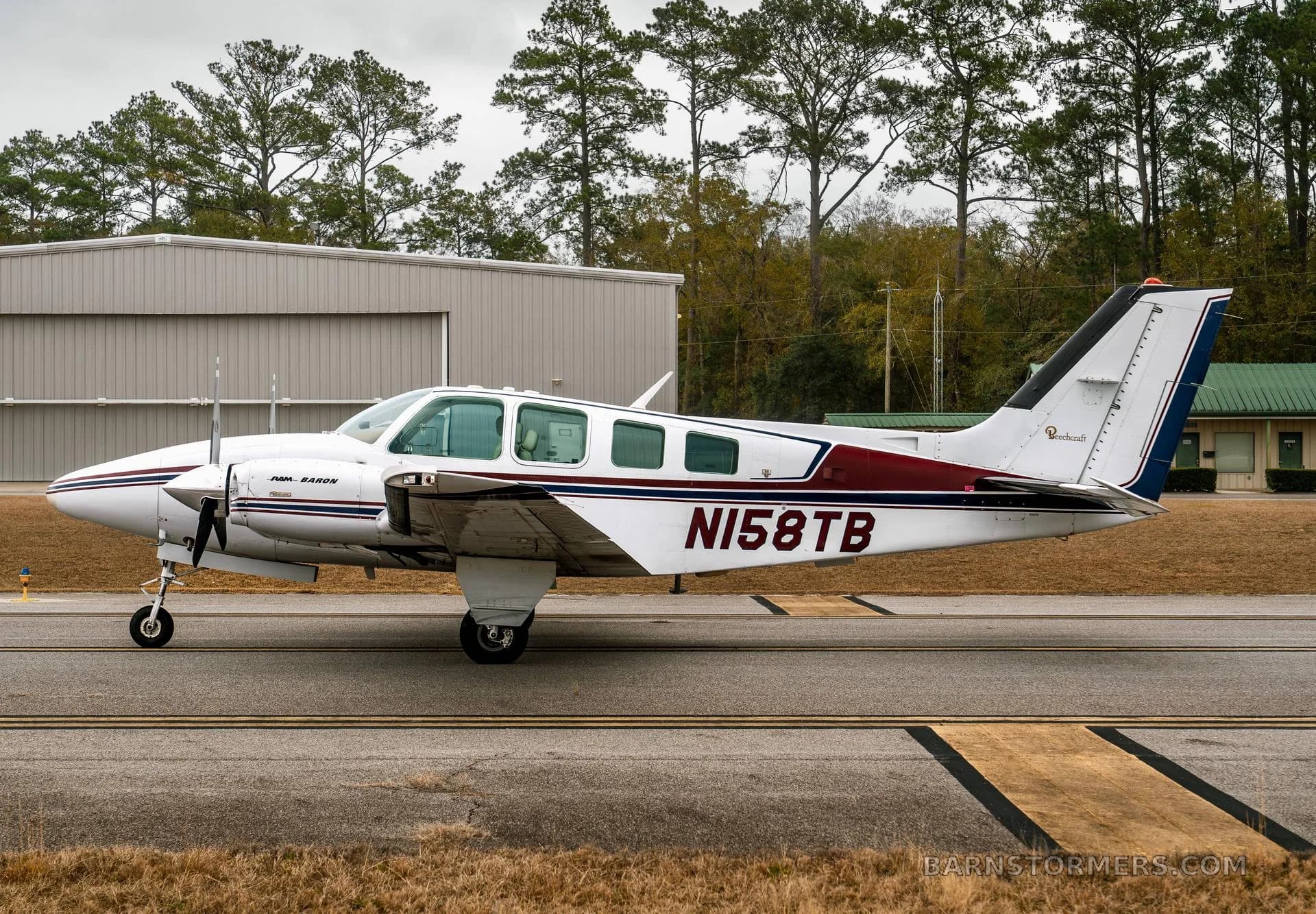A white twin-engine Beechcraft Baron N158TB with maroon and blue stripes is seen from the side on a taxiway, with a hangar and trees in the background.