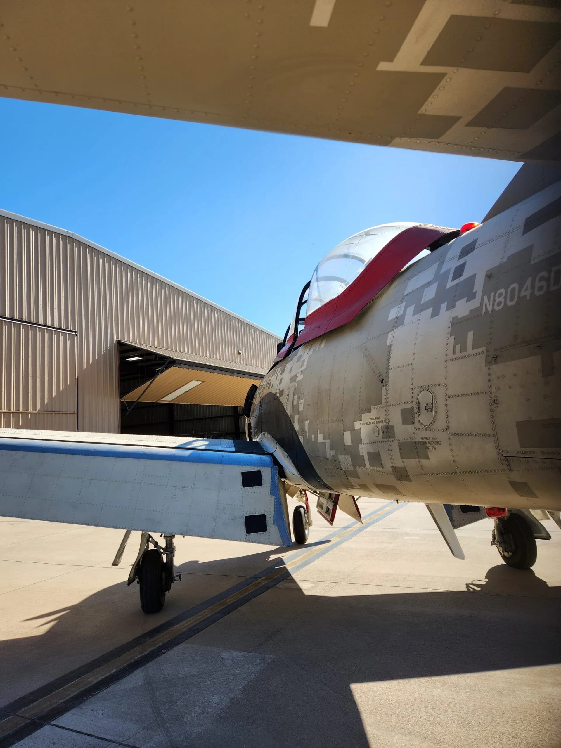 The tail section of a T-28 Trojan aircraft, showing its N-number N8046D and desert digital camouflage paint scheme, parked on an airfield.