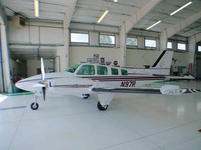 A white Beechcraft Baron N97R with red and black stripes is parked inside a brightly lit hangar.