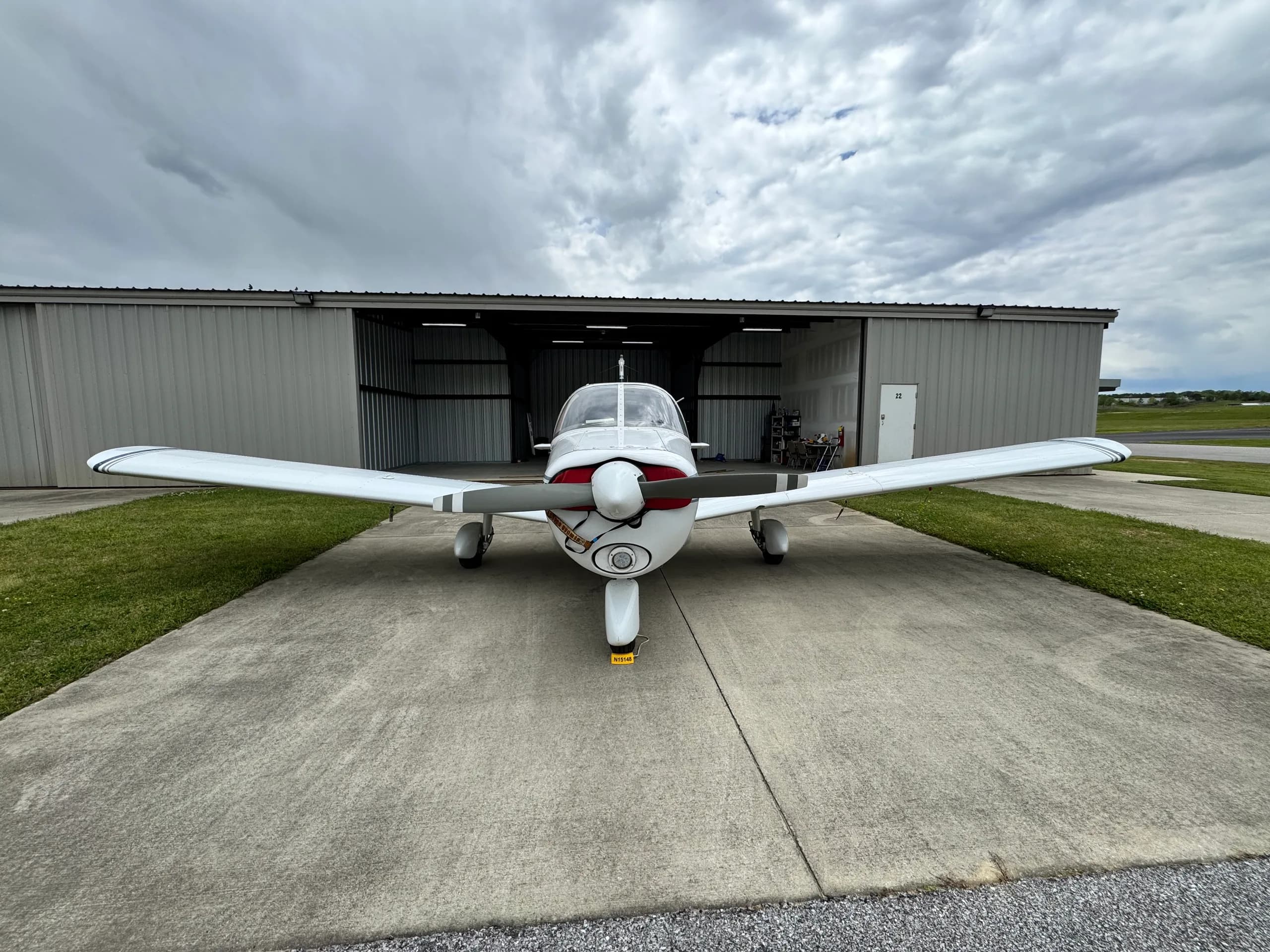 Head-on view of a white single-engine aircraft, N15148, with dark blue stripes, parked on concrete in front of an open hangar.