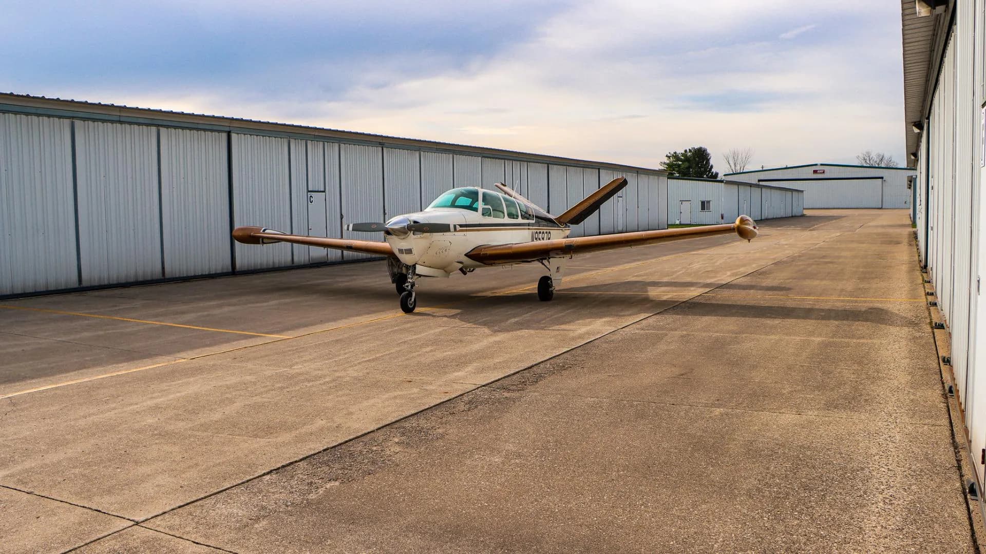 Distant front three-quarter view of a white and gold Beechcraft Bonanza parked on an airfield.
