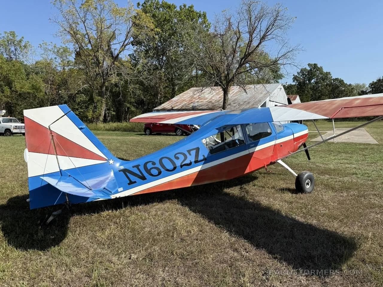 A red and blue high-wing aircraft with registration N602Z is parked on a grassy field next to a rustic building.