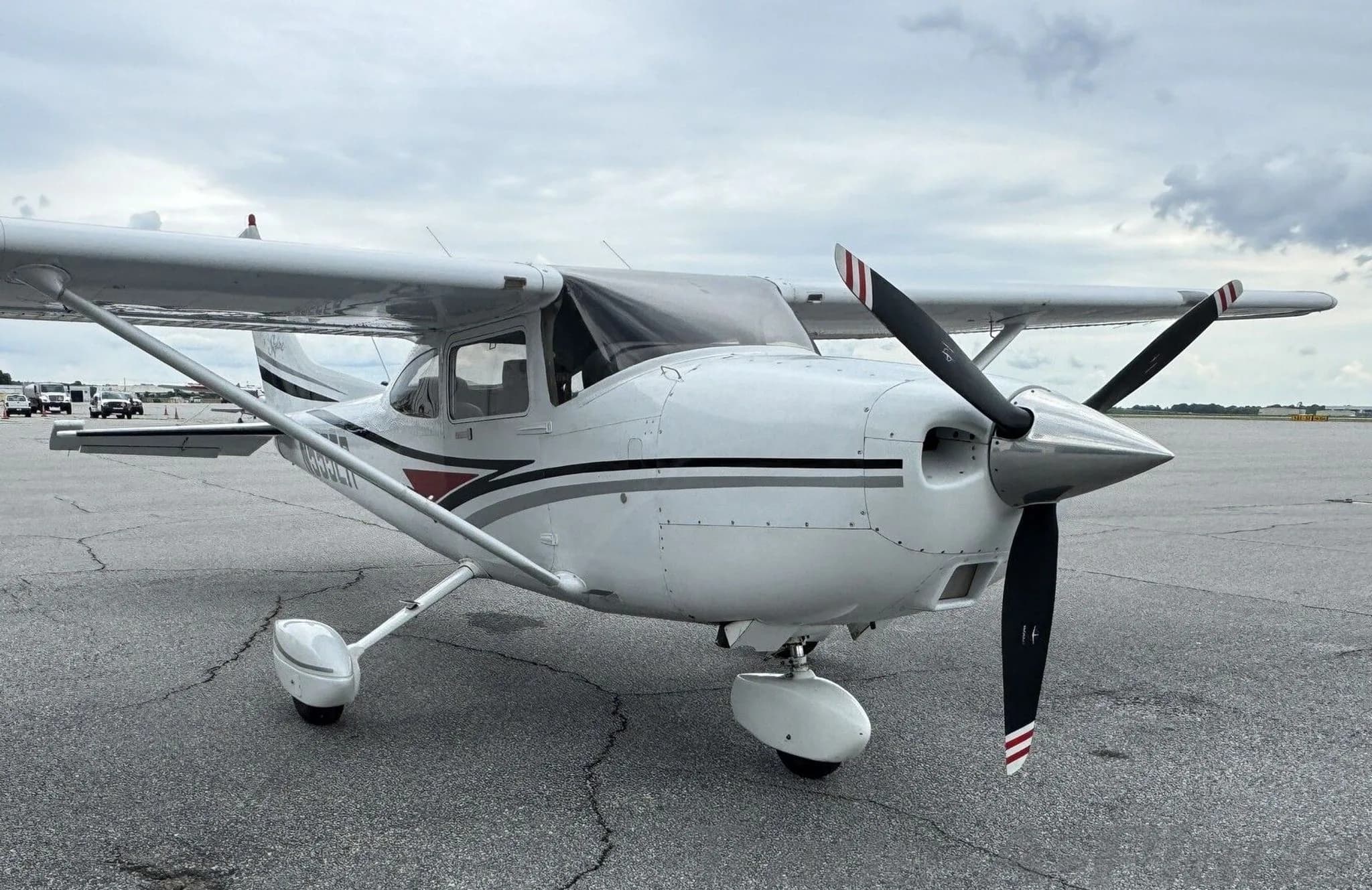 A front-quarter view of a white Cessna 182 Skylane with black, grey, and red stripes on an airfield.