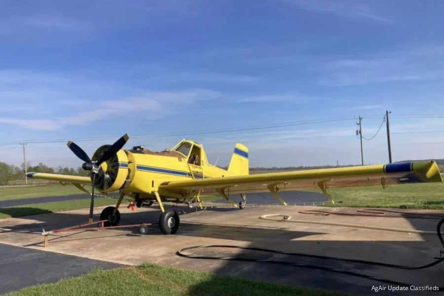 A yellow agricultural aircraft with blue stripes is parked on a tarmac under a clear blue sky.