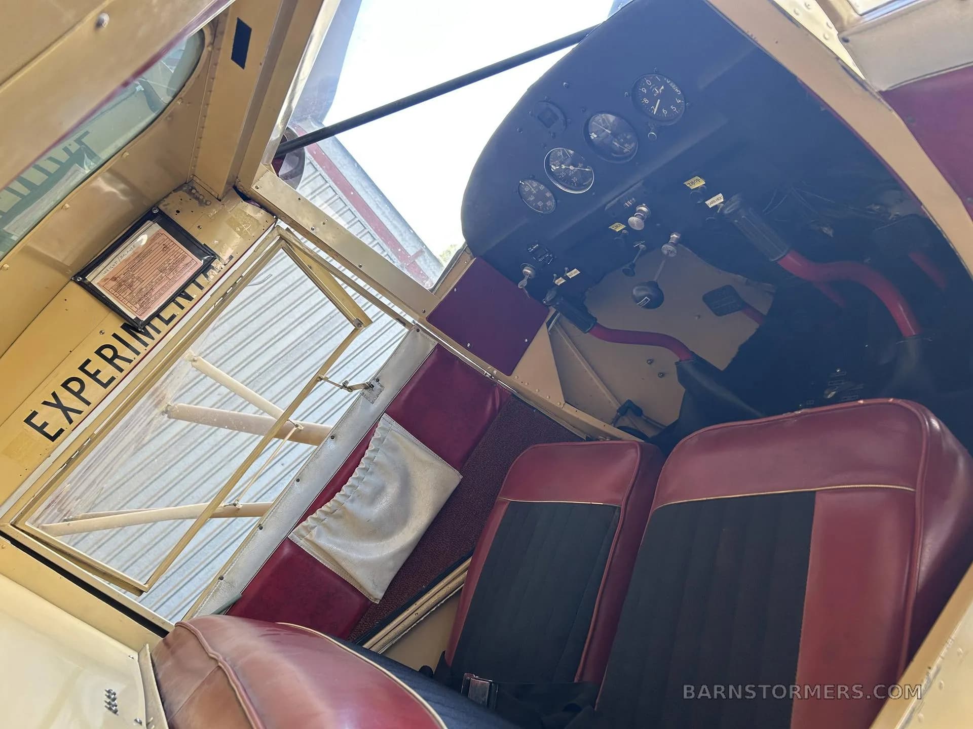 The interior of a vintage aircraft with red and black seats, showing the instrument panel and an open door with "EXPERIMENTAL" written on it.