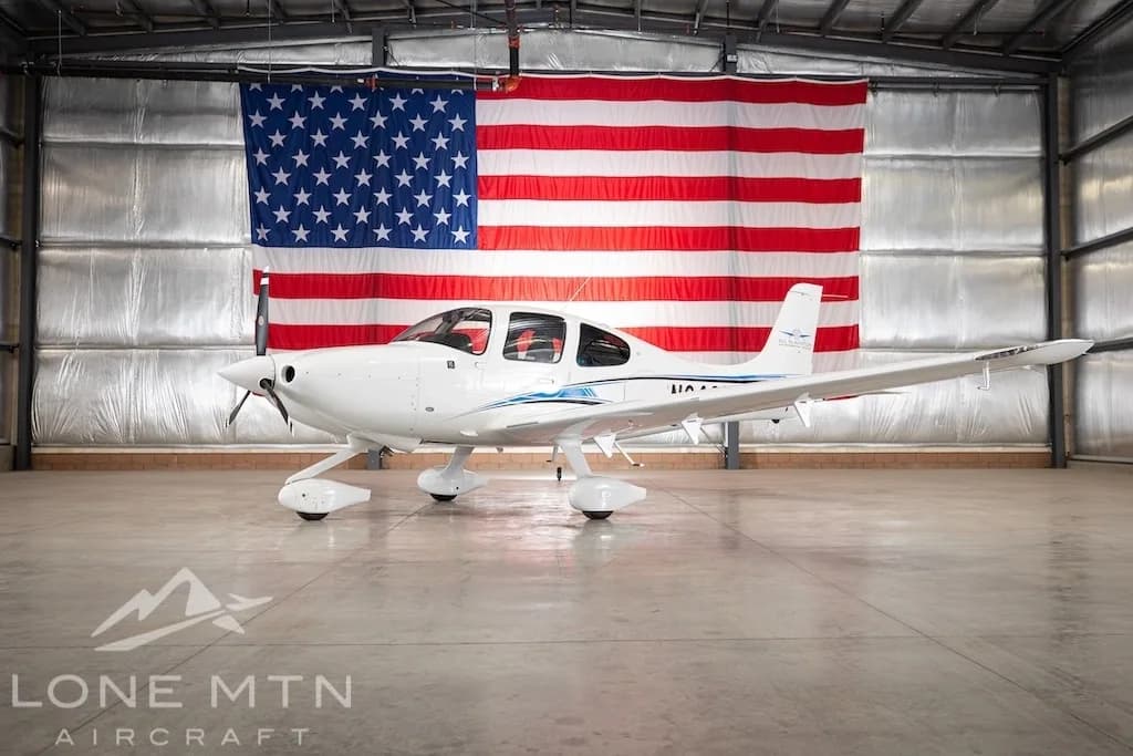 A white Cirrus SR22 aircraft with blue and black stripes, tail number N643LV, parked in a hangar with an American flag in the background.