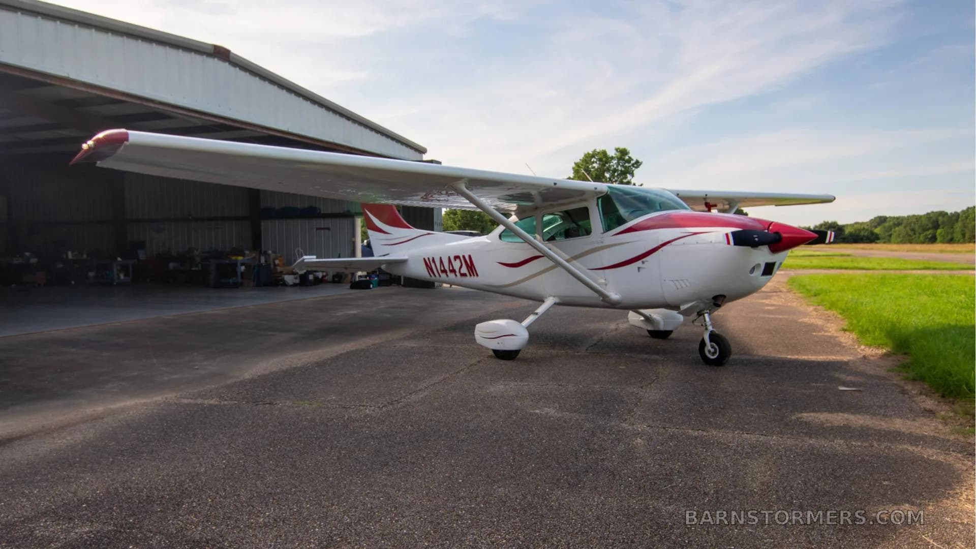 A white single-engine Cessna aircraft with red and gold stripes is parked on an asphalt ramp, viewed from the left side.