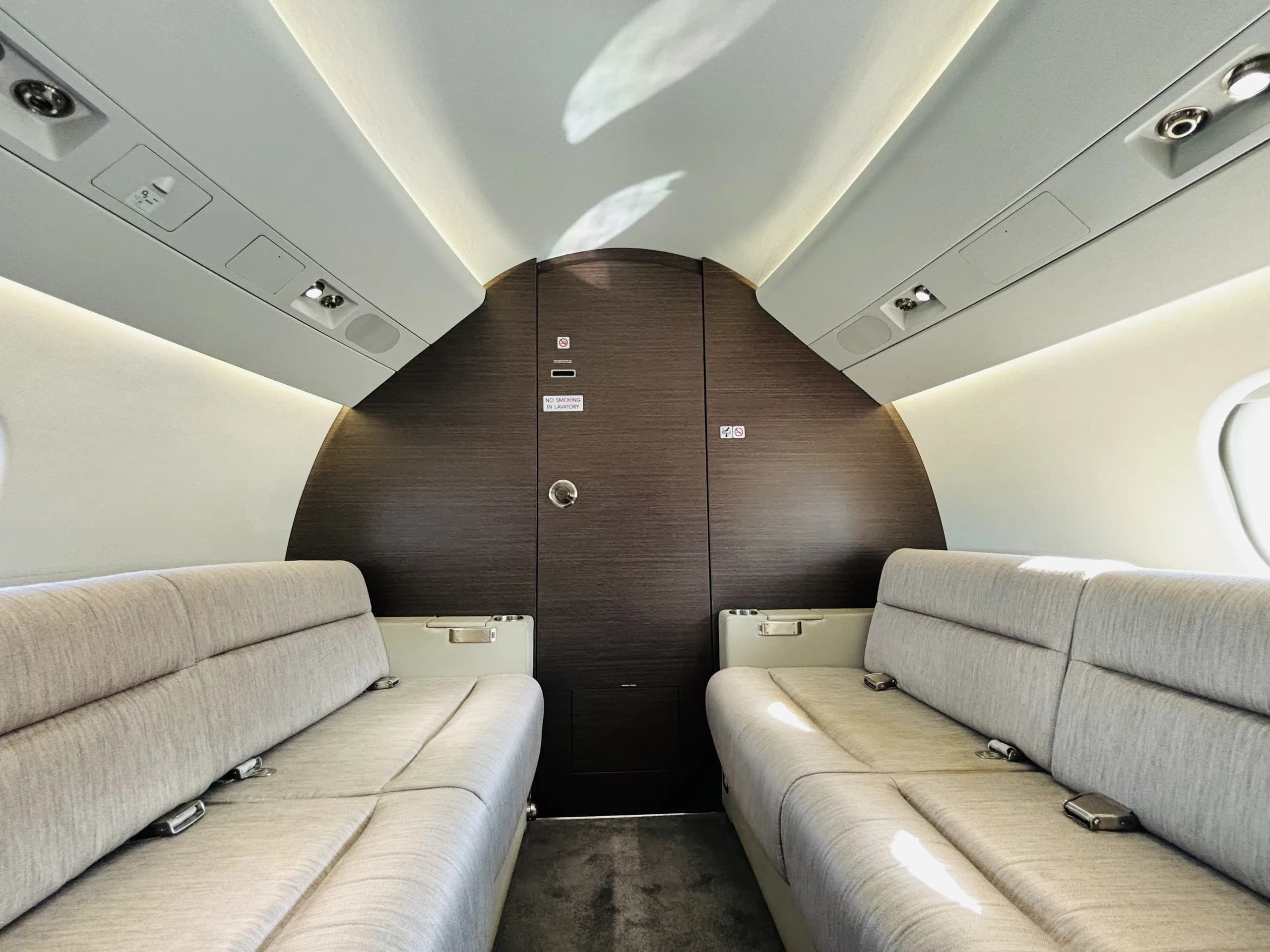 A view inside a private jet cabin showing two facing light grey fabric sofas and a dark wood bulkhead.