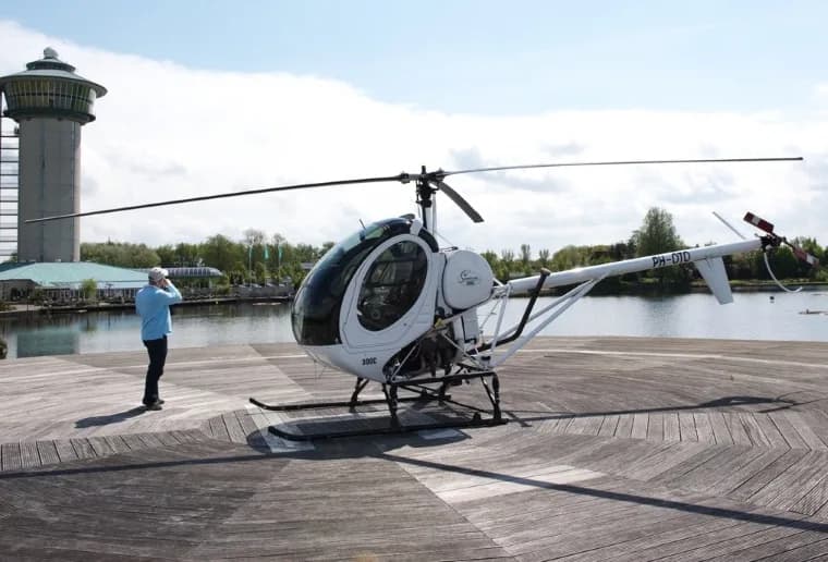 A white Schweizer 300C helicopter (PH-DTD) on a wooden helipad next to water, with a person and a control tower in the background.