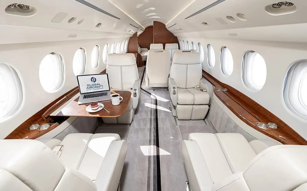 The luxurious interior cabin of a private jet with light-colored leather seats, a wooden table, and a laptop.