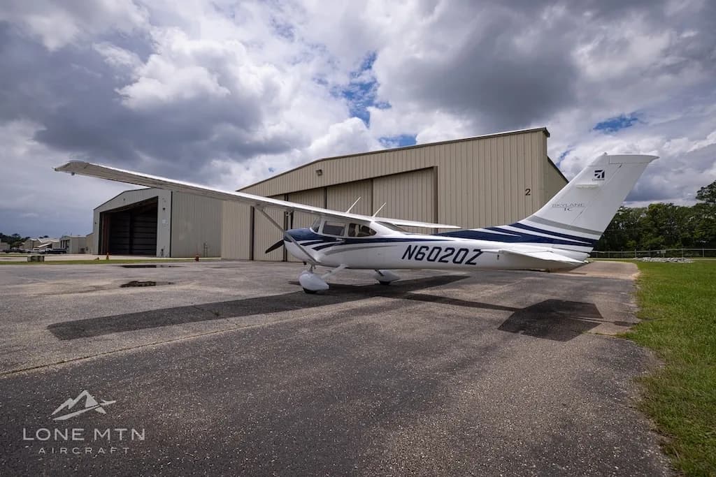 A right-side view of a white and blue Cessna Skylane TC (N6020Z) parked on an airfield next to a hangar.
