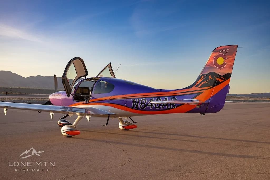 Three-quarter rear view of a purple and silver Cirrus SR22T with open doors, featuring a Colorado flag tail design, parked on a tarmac at sunset.