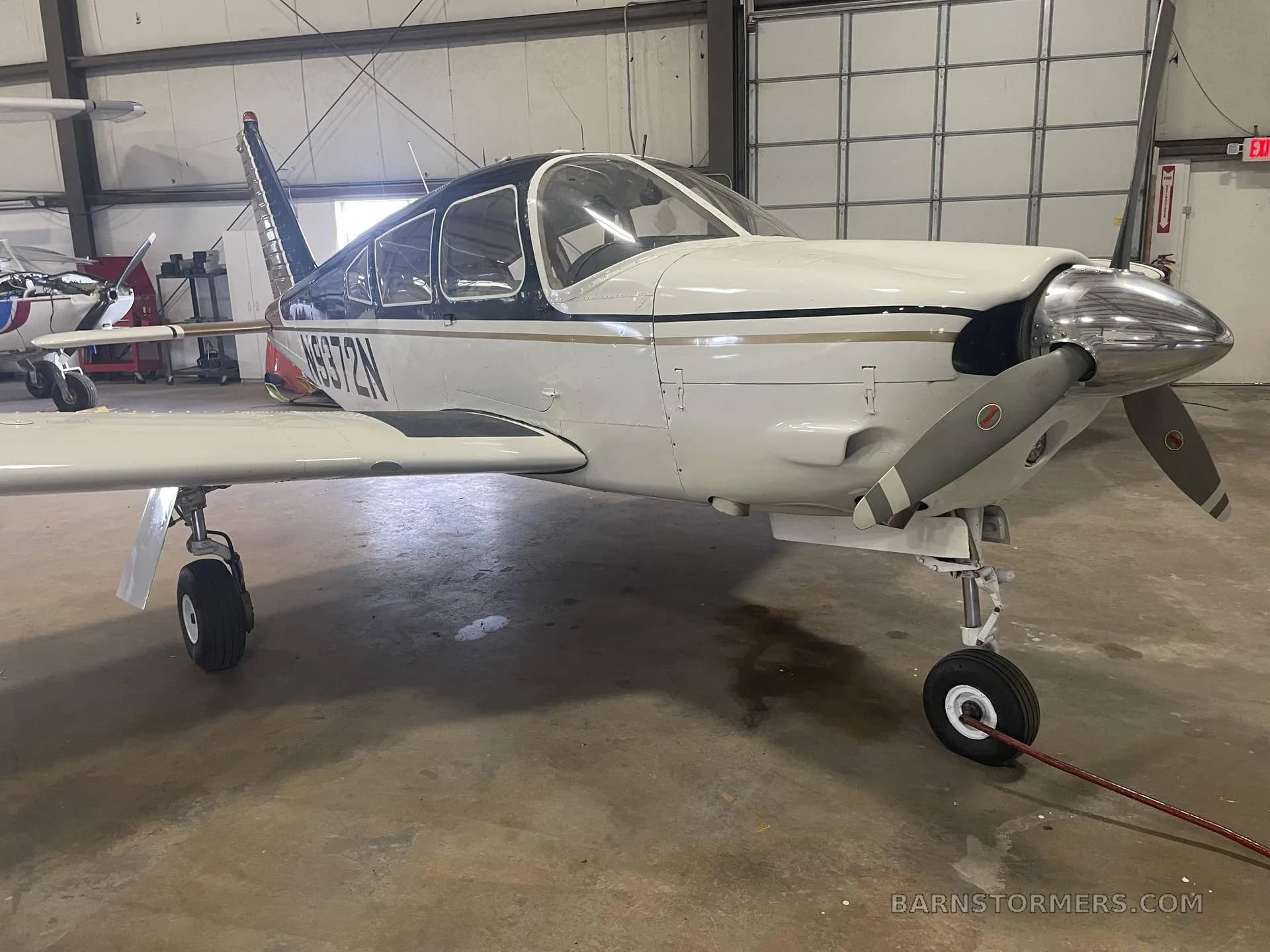 A white and black single-engine aircraft with a gold stripe, registration N9372N, parked in a hangar, viewed from the front-right.