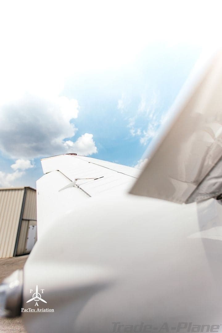 Upward view of the wing of a white aircraft with hangars in the background under a cloudy sky.