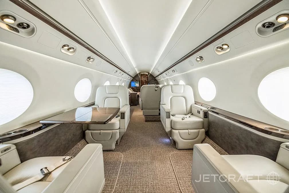 A luxurious aircraft cabin with light-colored leather seats and a wooden table, looking towards the rear.