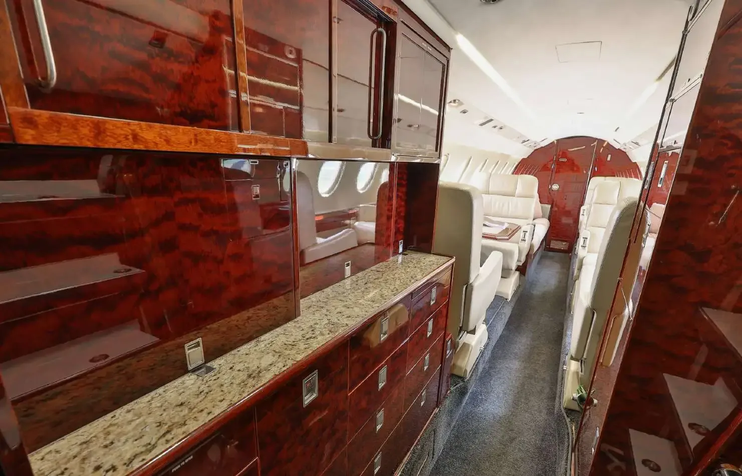 A view down the aircraft cabin aisle, showing a galley area with dark wood cabinetry and light-colored seats further back.