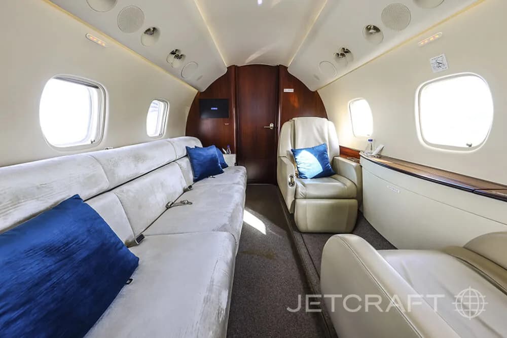 A luxurious private jet interior featuring a long beige sofa and a single beige armchair with blue accent pillows.