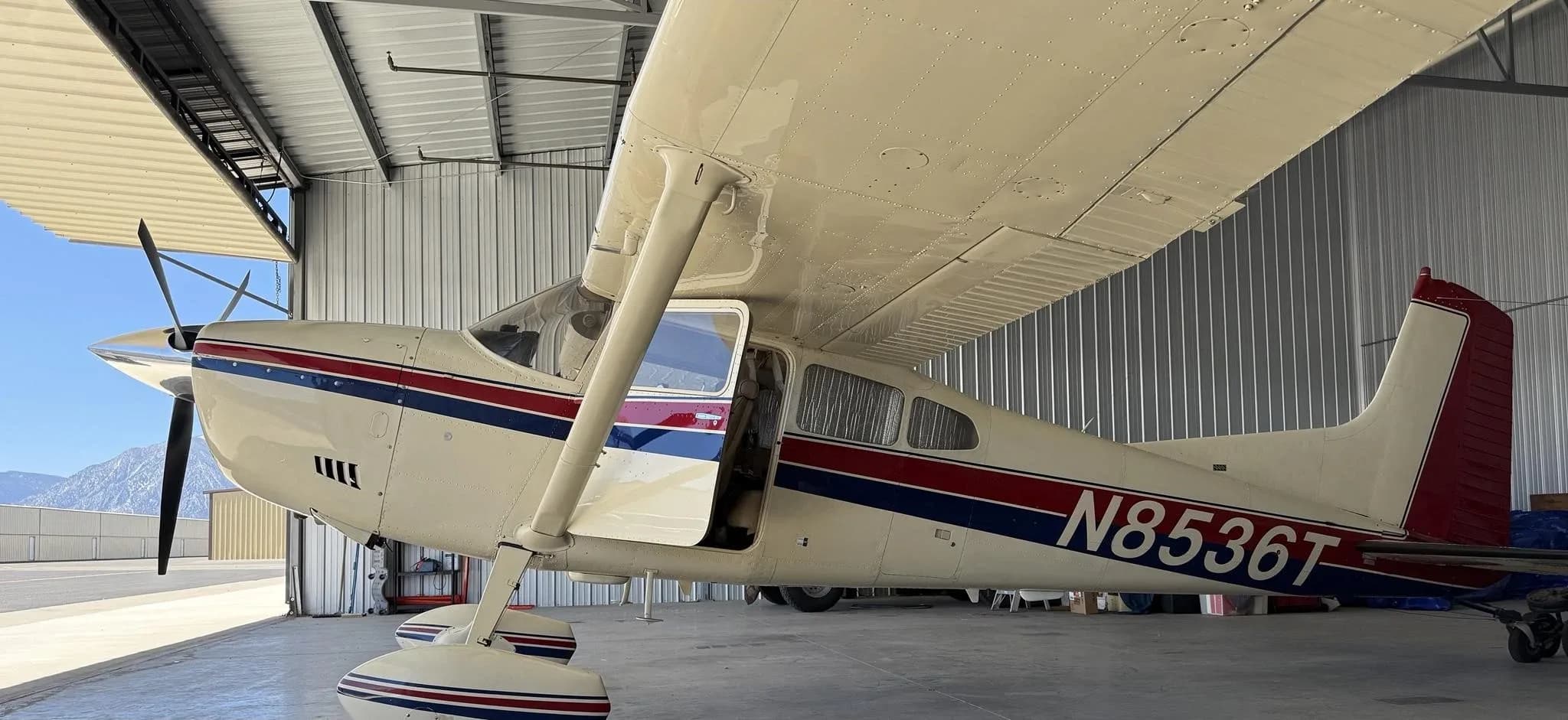 Side view of a white Cessna aircraft with red and blue stripes, registration N8536T, with its door open in a hangar.