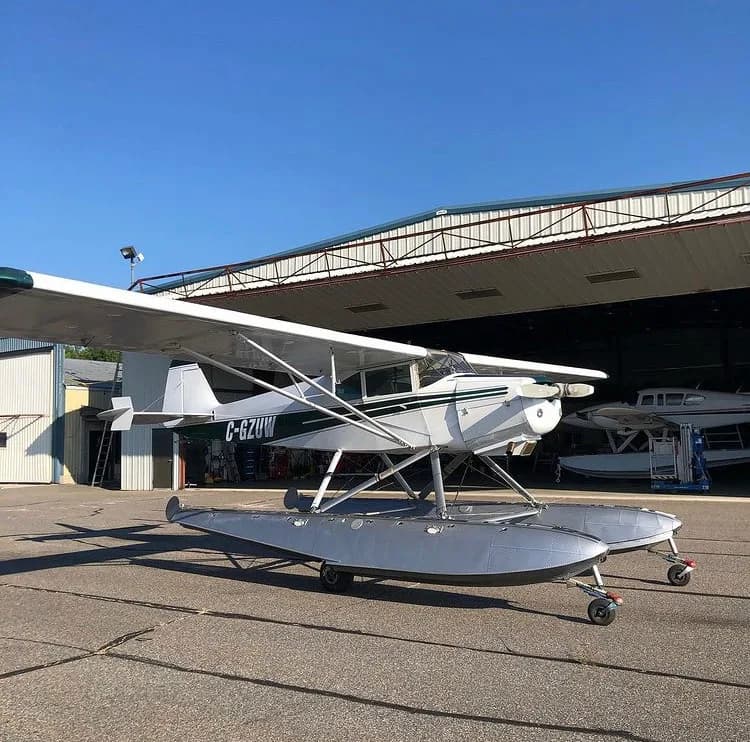 A white and green float plane, C-GZUW, is parked on the tarmac in front of a hangar.