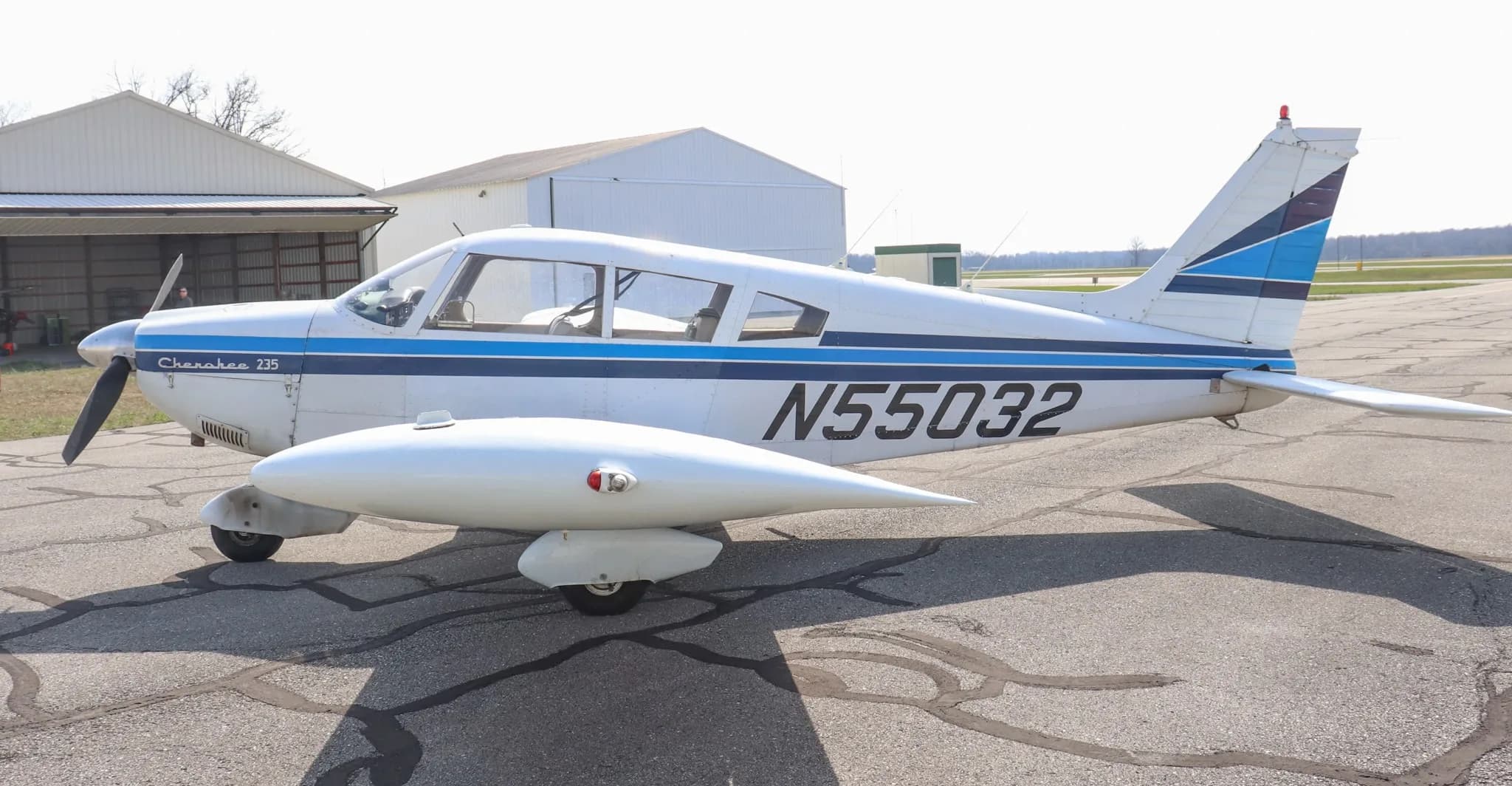 A side view of the white Piper Cherokee 235 aircraft with blue stripes and registration N55032, parked on an asphalt ramp.