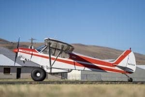 A Piper Super Cub aircraft with a red and white paint scheme is parked near a building.