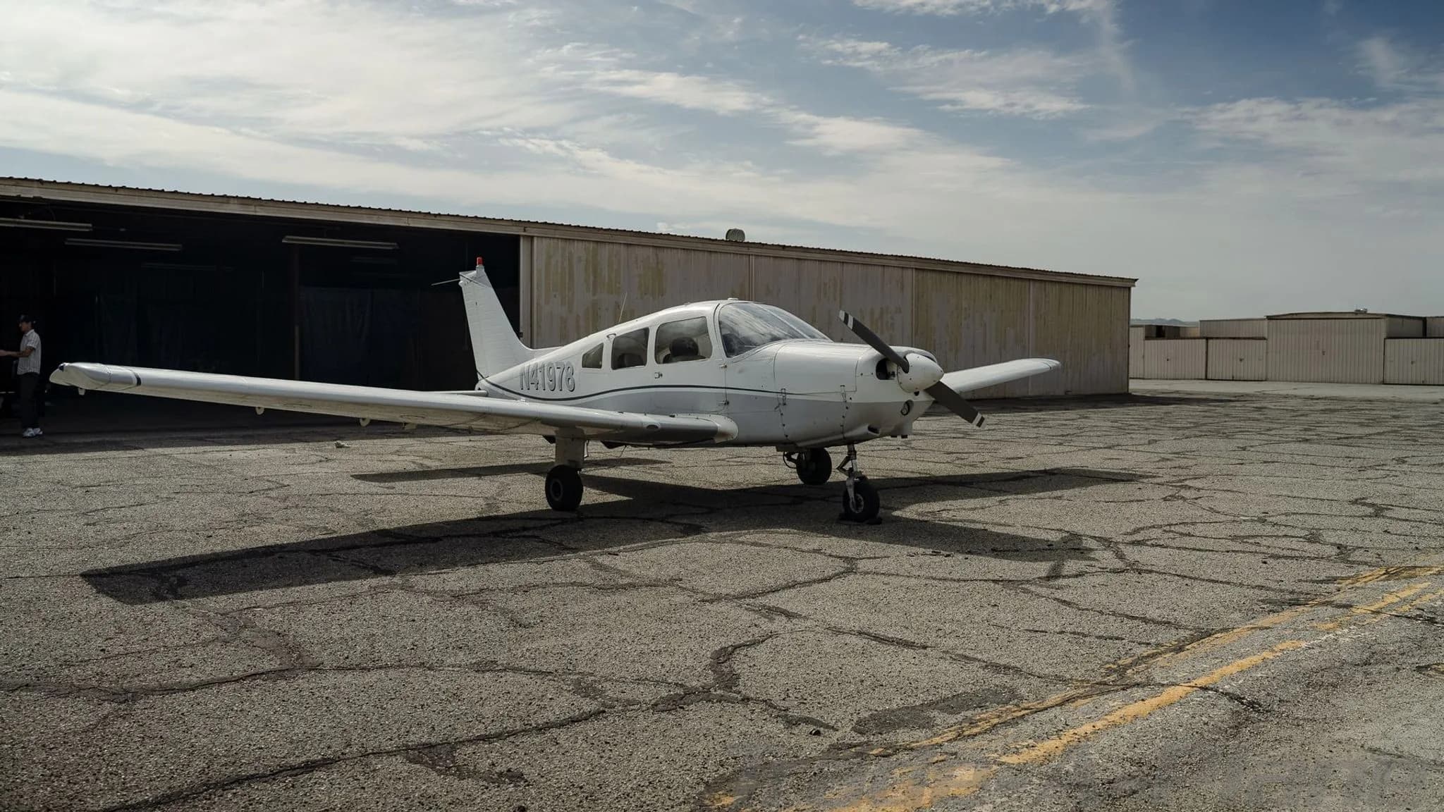 A white Piper Cherokee N41978 parked on a cracked tarmac with a hangar in the background.