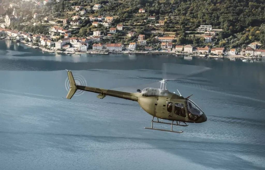 A green helicopter flies over water near a coastal town.