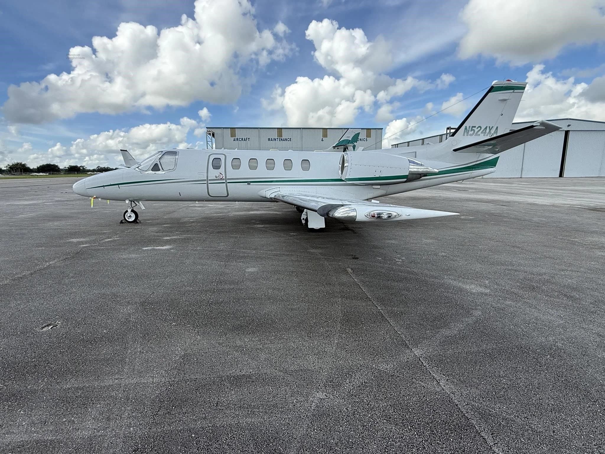 A white business jet N524XA with green stripes, parked on an airport tarmac with hangars in the background.