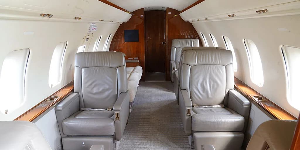 The interior cabin of a private jet features light grey leather seats, a beige carpet, and rich wooden paneling along the walls and a rear bulkhead.