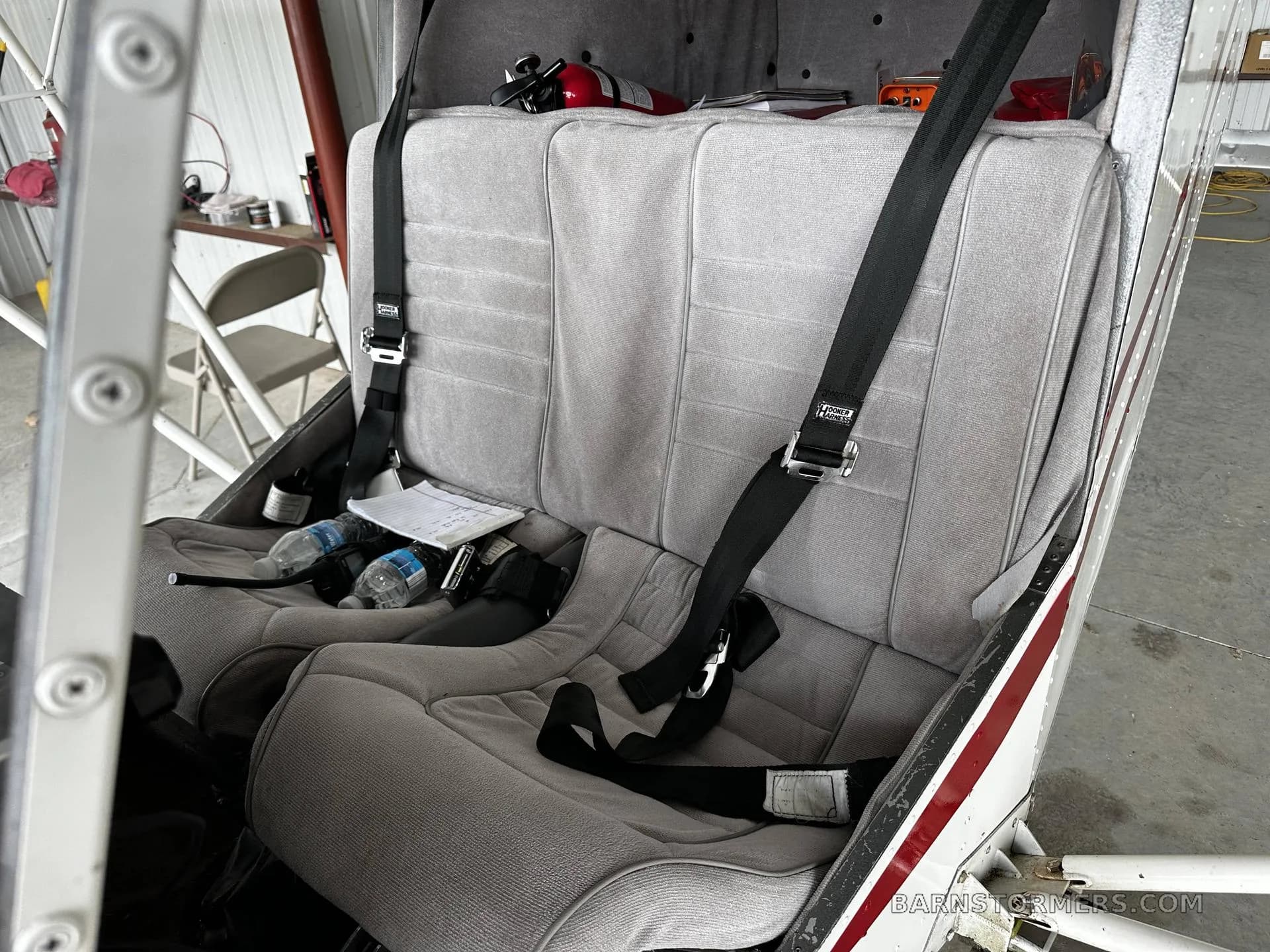 The light gray fabric seats of the aircraft cabin, equipped with black harnesses, with water bottles and papers on the seat.