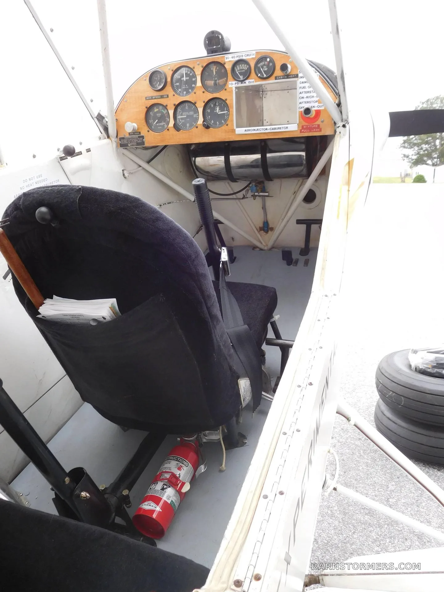 The cockpit of an experimental aircraft, showing the wooden instrument panel, a black fabric seat, and a fire extinguisher.