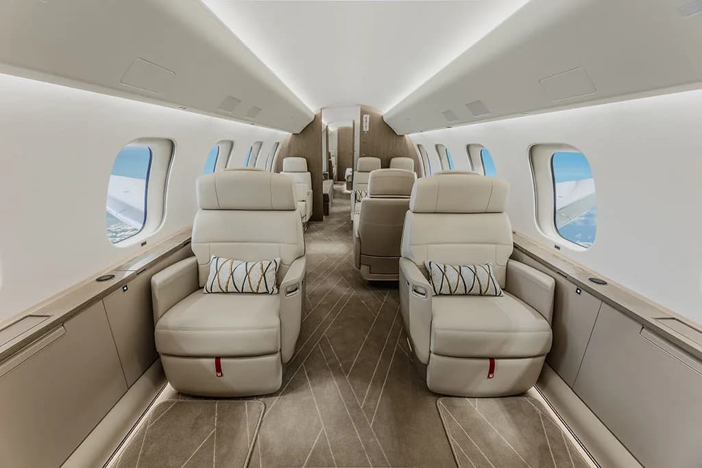A view inside a private jet cabin showing two rows of beige leather seats with decorative pillows.