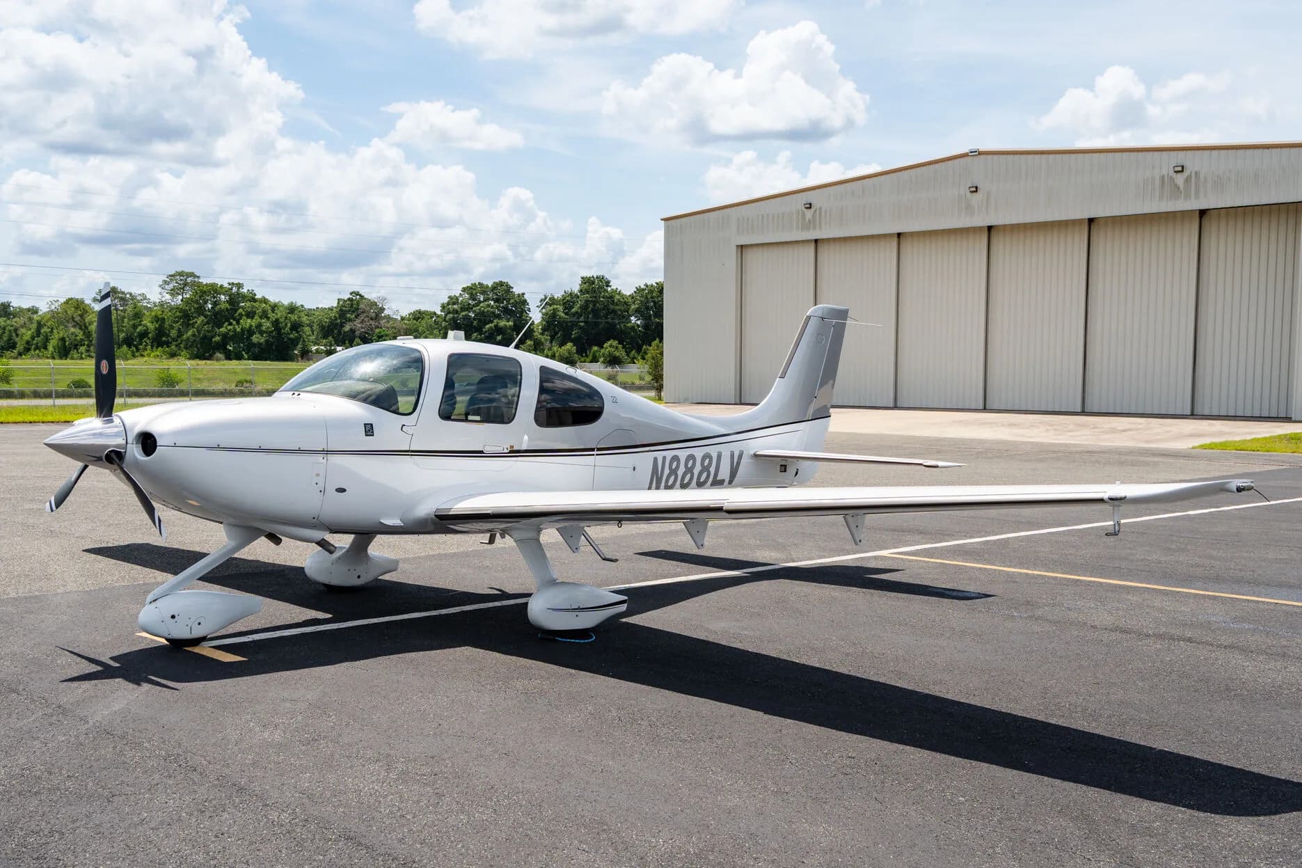 Three-quarter front view of a silver Cirrus SR22 aircraft (N888LV) parked on an airfield next to a hangar.