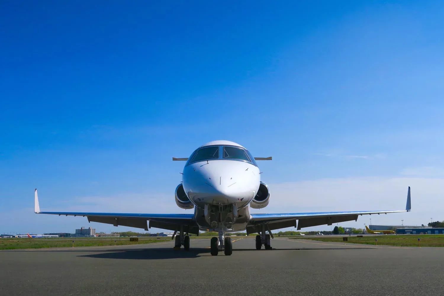 A white business jet with two engines, viewed head-on on an airport tarmac under a clear blue sky.
