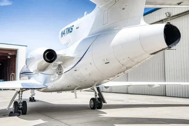 A low-angle rear view of a white Dassault Falcon 900EX business jet with blue stripes, registration N47NS, showing the three rear-mounted engines.