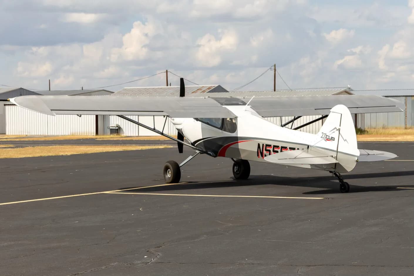 A side view of the white and grey X-Cub aircraft N555ZM with red accents, parked on an asphalt runway with hangars in the background.