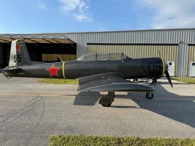 A dark gray Yak-52 aircraft with red stars and yellow accents parked on an asphalt ramp next to hangars.