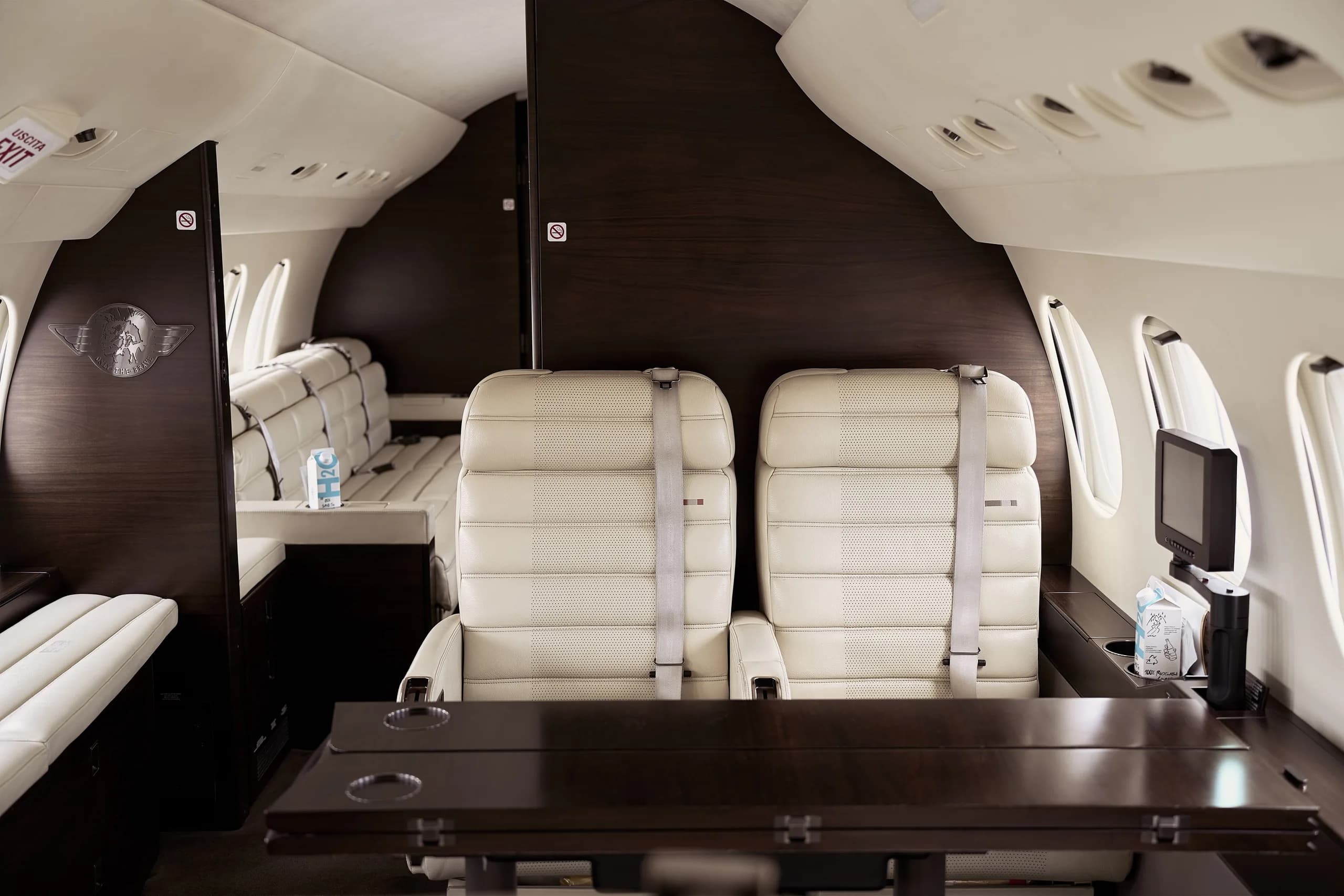 A close-up view of two cream leather aircraft seats with a dark wooden partition and table in a private jet cabin.