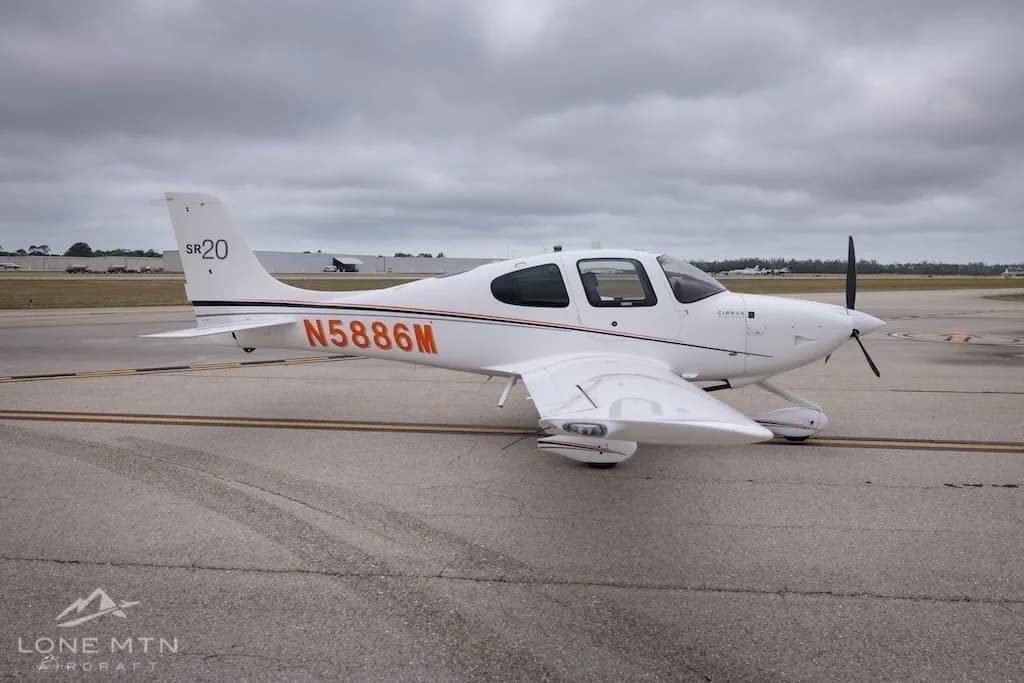 A three-quarter front view of a white Cirrus SR20 aircraft with orange and black stripes, registration N5886M, on an airfield.