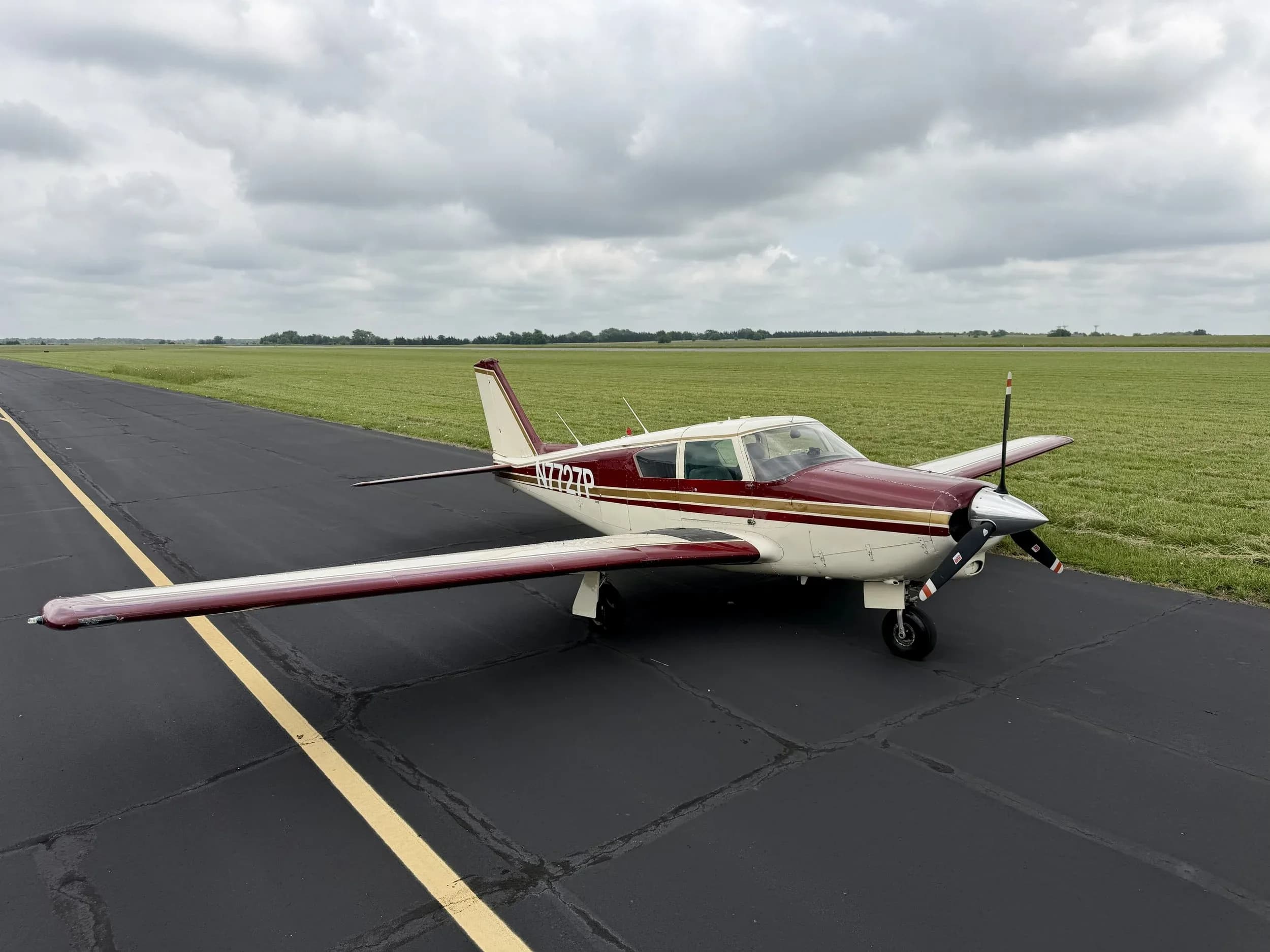 A maroon and white Piper Comanche N7727P parked on an asphalt runway with green fields in the background.